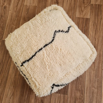 A top-down view of a square, handmade Beni Ourain style pouf. It features a high-pile shag texture in creamy ivory wool, with abstract black lines forming a minimalist pattern