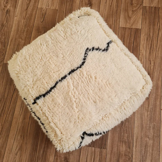A top-down view of a square, handmade Beni Ourain style pouf. It features a high-pile shag texture in creamy ivory wool, with abstract black lines forming a minimalist pattern