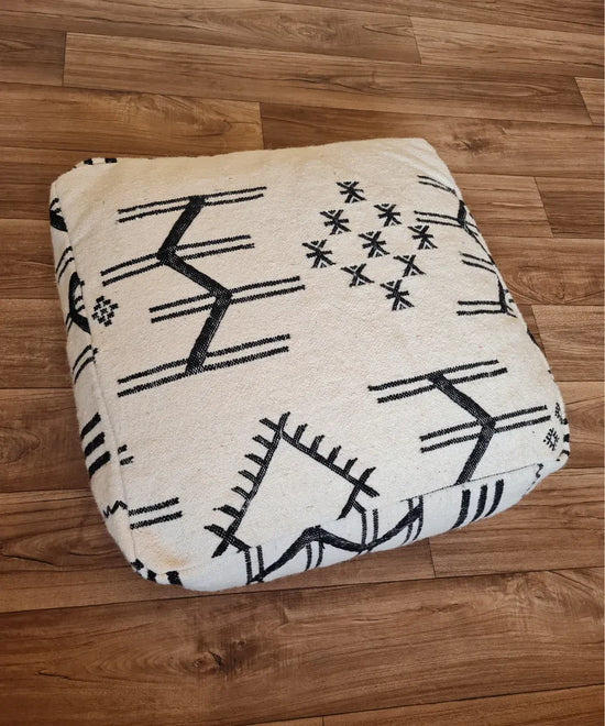 A top-down view of a square, handmade floor cushion made from a modern Moroccan woven rug. The cream wool base is decorated with bold, black geometric and tribal Berber symbols