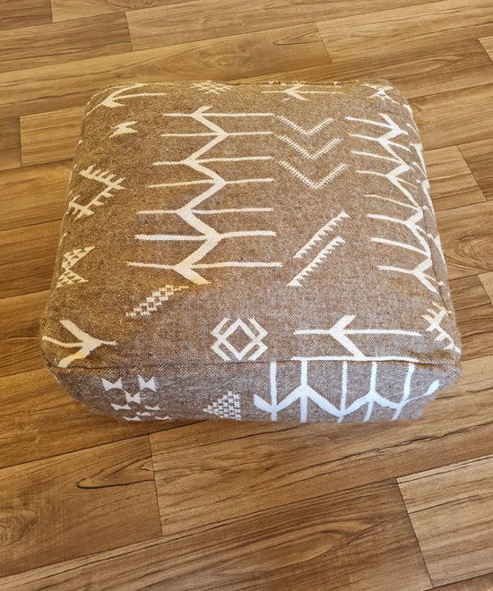 A top-down view of a square, handmade floor cushion made from a modern Moroccan woven rug. The earthy brown fabric base is decorated with bold, graphic white Berber-inspired symbols