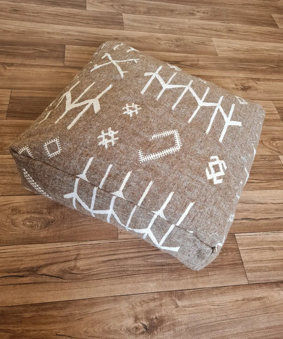 A top-down view of a square, handmade floor cushion made from a modern Moroccan woven rug. The earthy brown fabric base is decorated with bold, graphic white Berber-inspired symbols