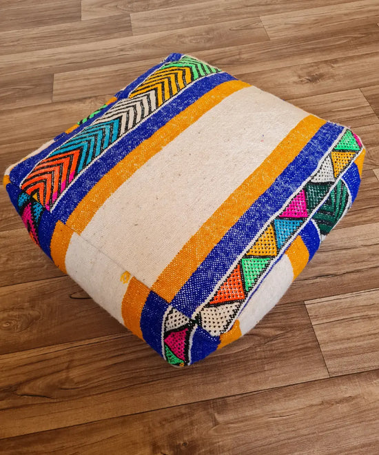 A top-down view of a square, handmade floor cushion made from a modern Moroccan Kilim. The cream wool base is decorated with bold stripes in royal blue and yellow, accented with colorful geometric patterns