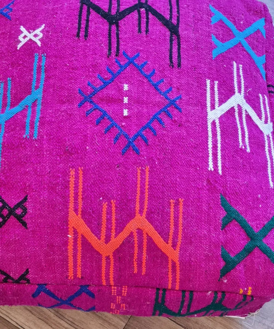A top-down view of a square, handmade floor cushion made from modern Moroccan woven wool. The vibrant fuchsia base is decorated with colorful embroidered Berber symbols in blue, orange, black, and white