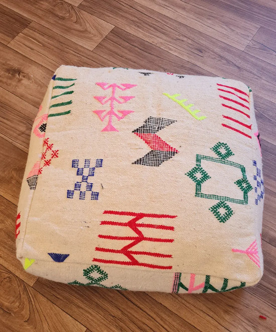 A top-down view of a square, handmade floor cushion made from a modern Moroccan Kilim. The cream wool base is decorated with colorful embroidered Berber symbols in neon pink, yellow, green, and red