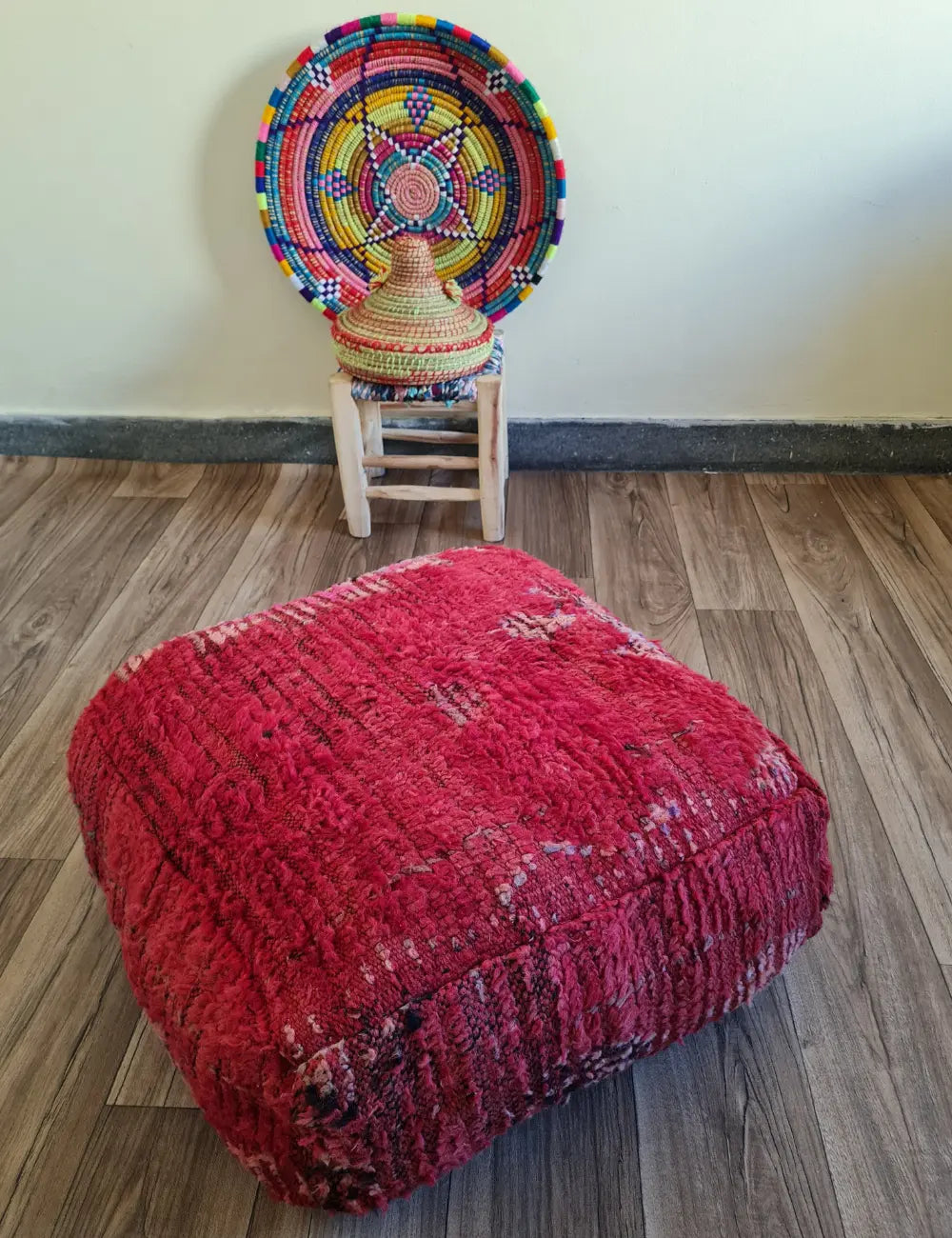 A square vintage rug floor cushion in shades of pink and red on a wood floor. In the background is a small wooden stool with a colorful Moroccan basket
