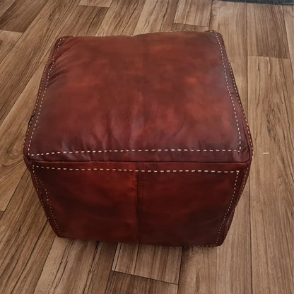 A square brown leather pouf on a wood floor. In the background is a small wooden stool with a colorful Moroccan basket and plate
