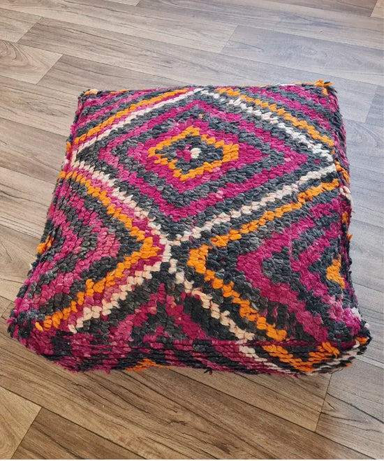 A top-down view of a square, handmade floor cushion made from a vintage Kilim rag rug. It has a high-pile shag texture with a geometric diamond pattern in magenta, orange, charcoal, and white