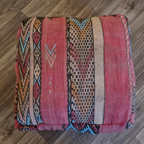 A top-down view of a square, handmade floor cushion made from a vintage Moroccan Kilim rug. It has a flat-weave texture with tribal geometric stripes in dusty rose, red, blue, and orange