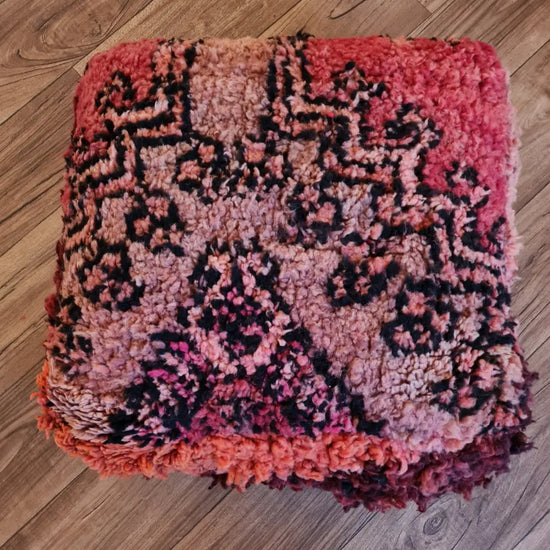 A top-down view of a square, handmade floor cushion made from a vintage Moroccan wool shag rug. It has a high-pile texture with abstract tribal patterns in deep pink, red, orange, and black