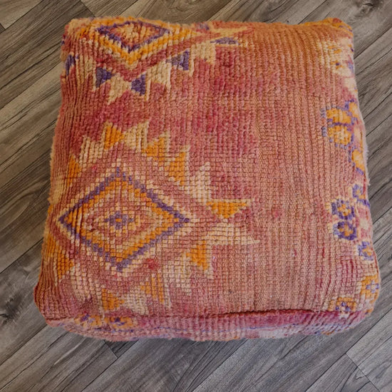 A top-down view of a square, handmade floor cushion made from a vintage Moroccan wool rug. It has a low-pile texture with sun-faded tribal patterns in shades of dusty rose, orange, and purple
