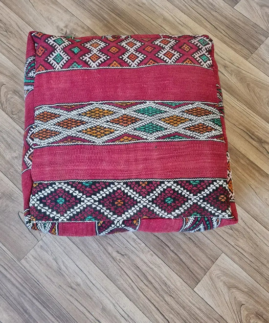 A top-down view of a square, handmade floor cushion made from a vintage Moroccan Kilim rug. It has a flat-weave texture with intricate geometric Berber patterns in red, green, yellow, orange, and white