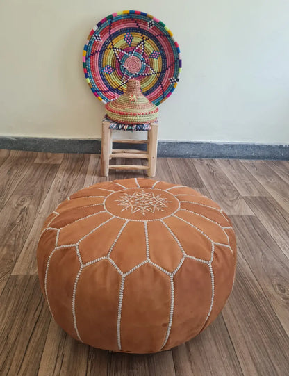 A tan suede Moroccan pouf on a wood floor. In the background is a small wooden stool with a colorful Moroccan basket and plate