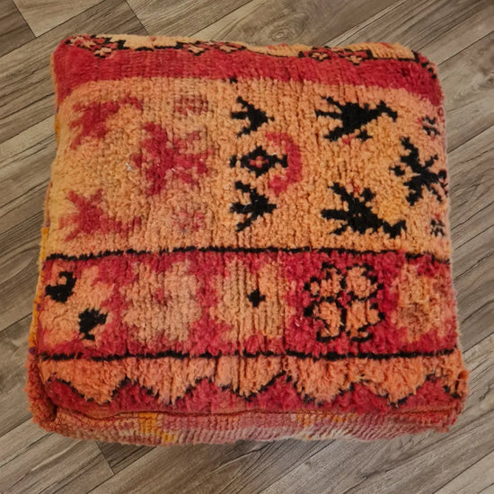 A top-down view of a square, handmade Moroccan floor cushion made from a vintage Boucherouite rug. It has a high-pile shag texture with tribal patterns in red, orange, pink, and black