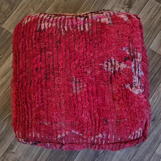 A top-down view of a square, handmade Moroccan floor cushion made from a vintage Boucherouite rug. It has a high-pile shag texture in vibrant shades of pink and red