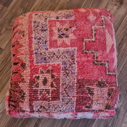 A top-down view of a square, handmade floor cushion made from a vintage Moroccan wool rug. It features a plush texture with Berber geometric patterns in shades of magenta, pink, and deep red