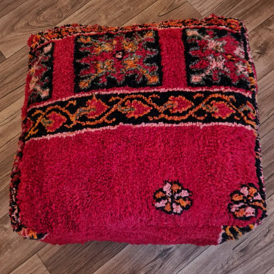 A top-down view of a square, handmade floor cushion made from a vintage Moroccan wool shag rug. It features a deep red high-pile texture with a wide band of black and orange tribal patterns
