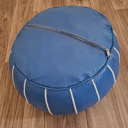 The underside of a blue leather pouf, showing the large zipper used for stuffing, set against a dark wood floor