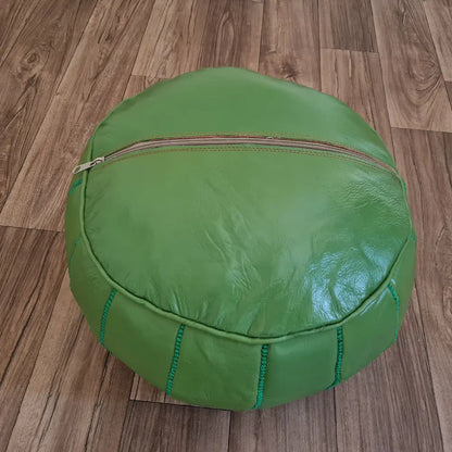 The bottom of a green leather pouf, showing the large zipper used for stuffing, set against a dark wood floor