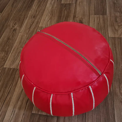 The underside of a red leather pouf, showing the large zipper used for stuffing, set against a dark wood floor