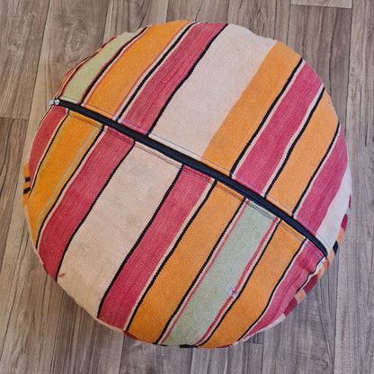 The bottom of a round Kilim pouf, showing a striped pattern in pink, orange, and cream, with a large black zipper for stuffing