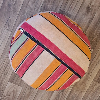 The bottom of a round Kilim pouf, showing a striped pattern in pink, orange, and cream, with a large black zipper for stuffing
