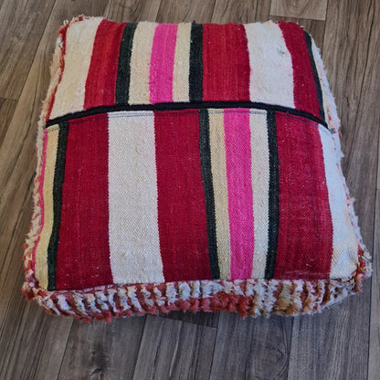 The bottom of a square Boucherouite pouf, showing a striped flat-weave pattern in shades of pink, red, and cream, with a large black zipper for stuffing